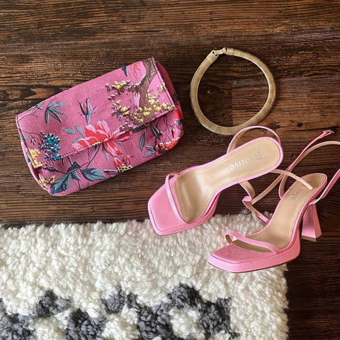 A pink pair of heels and a pink floral clutch are tossed on a white and grey fluffy rug.