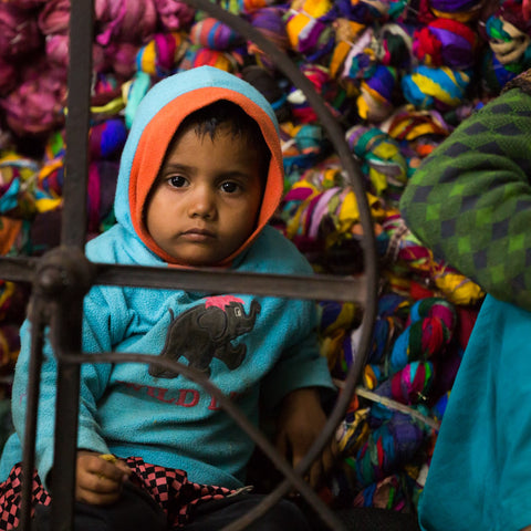 A little boy wearing a bright blue hoodie sits beside his mother behind her spinning wheel. Behind him is a wall of colorful ribbon yarn.
