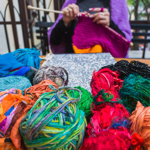 A woman wearing a purple sweater is sitting at a table knitting. In front of her is a bunch of Darn Good Yarn - all reclaimed, recycled, handmade, and sustainable yarn