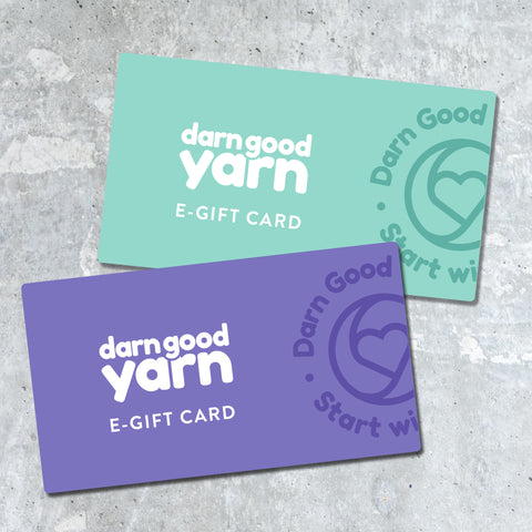 Two darn good yarn gift cards, one mint green and the other a soft purple, are laying on a soft gray marble tabletop.