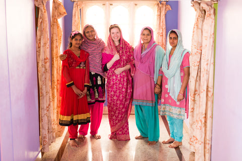 This is a picture of our founder and CEO, Nicole, along with some of our artisans. They are each wearing different colored saris in a brightly lit room.