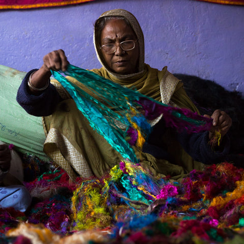 An Indian woman wearing glasses is sitting on the floor, tearing multicolored recycled sari strips.