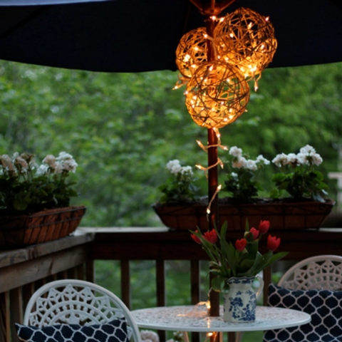 On a deck, a pair of hemp ball lanterns are hung up underneath a dark blue umbrella at a white iron table.