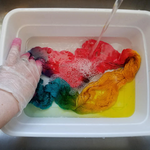 A gloved hand is holding down a loose skein of rainbow dyed yarn in a white plastic bin. At the upper right side of the bin, a stream of clear water is being poured into the container. The yarn is starting to become yellow as the extra dye bleeds out into the water.