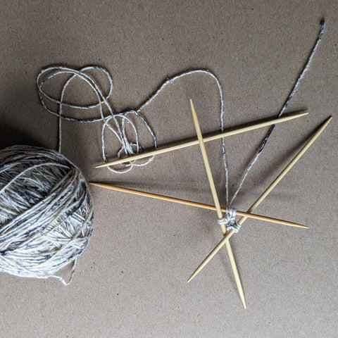 A ball of white and gray speckled yarn is resting on a wooden board. Next to the ball of yarn are two sets of double pointed needles.