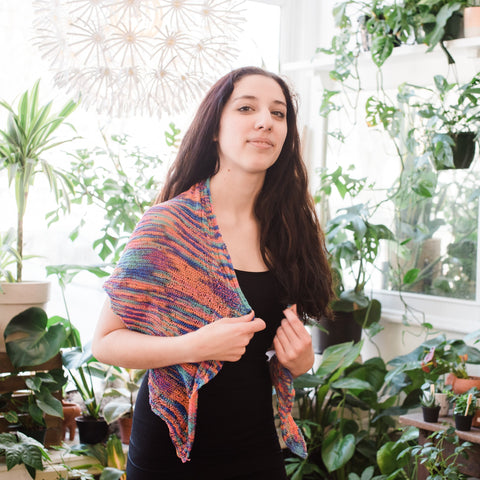 A woman with long brown hair, a black tank top, and wearing a brightly colored lace weight shawl around her shoulders, is posing in a greenhouse.