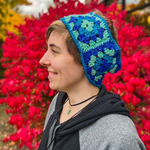 A person with short brown hair is facing sideways, wearing a multi-colored blue head warmer, a black choker, and a black and grey hoodie.