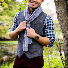 A man standing out in the woods, wearing  a checkered shirt, a grey vest, and a thin pastel purple/grey scarf.