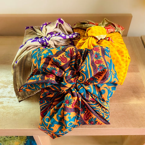 Three different boxes, which have been wrapped in different colored (yellow, tan, orange/teal) furoshiki reusable gift wrapping.