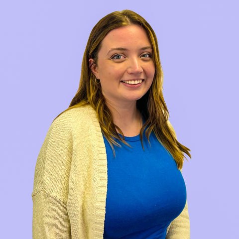 Close up of the author, Gabby, who's wearing a bright blue top and a soft yellow cardigan. She's smiling at the camera and sitting in front of a light purple wall.