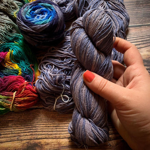 A hand holding a skein of dark purplish grey mulberry lace weight silk yarn.