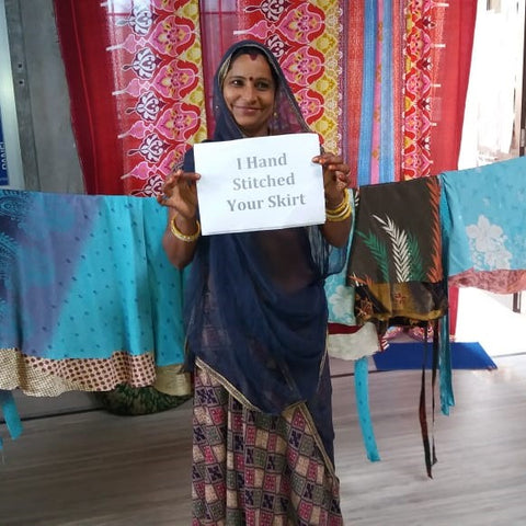 An artisan wearing a deep purple sari is standing in front of a close-line of finished skirts. She's smiling and holding a sign that says "I hand stitched your skirts: