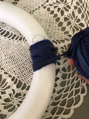 Woman's hand wrapping blue ribbon around a foam white wreath on top of a white crochet blanket