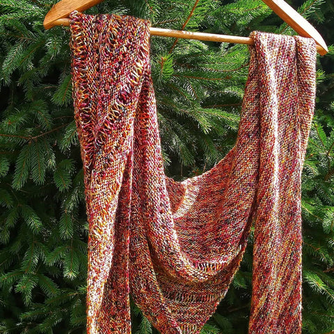 An orange and white shawl is hanging off a wooden hangar, in front of an evergreen tree.