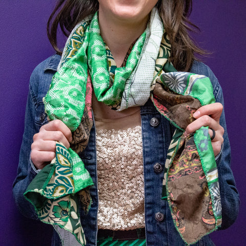 A woman from the chin down, wearing a golden shirt, a jean jacket, and a green scarf