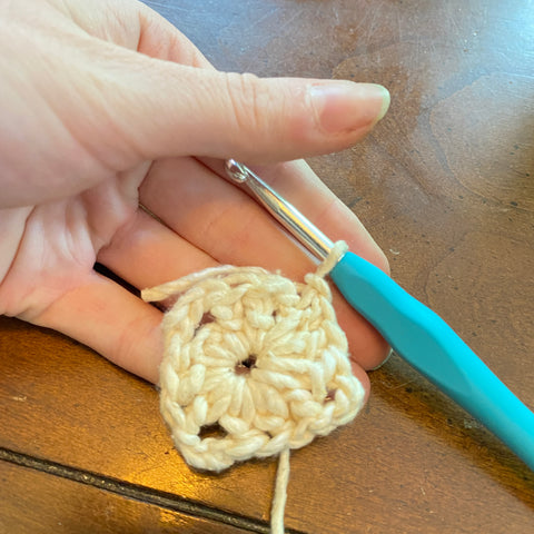 A pale hand is holding the finished center of a granny square, made of white worsted weight yarn. A light blue crochet hook is still attached to the yarn.