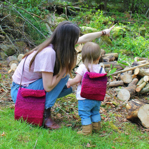 Back view of a woman and baby wearing matching pink crochet bags and standing on grass next to a pile of wooden logs