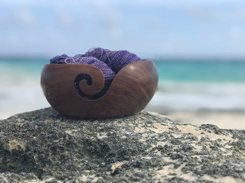 Wooden yarn bowl full of purple sparkly yarn sitting on a rock in front of an ocean