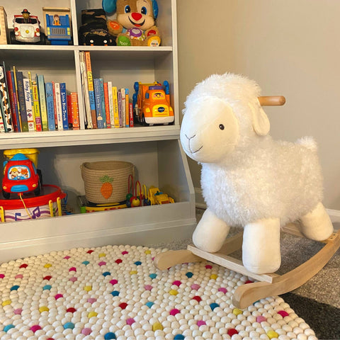 A white and rainbow dotted felt ball rug is laid out in a babies room. To the right of the screen is a rocking sheep and to the left is a bookshelf filled with books and toys.