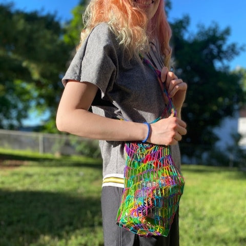 A person with long blonde hair has a mesh bag made of rainbow lace weight over their shoulder.