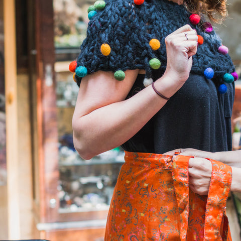 A person wearing an orange wrap skirt, a black top, and a black and multicolored felt ball shrug