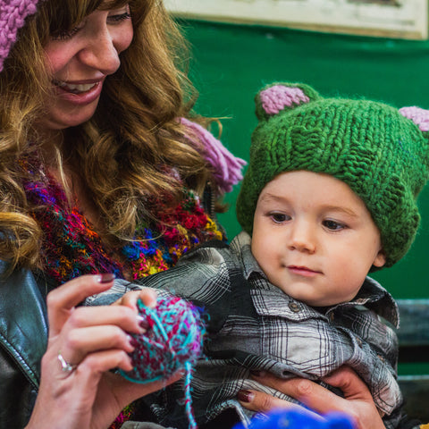 A woman is holding her child, who is reaching for a colorful ball of yarn.