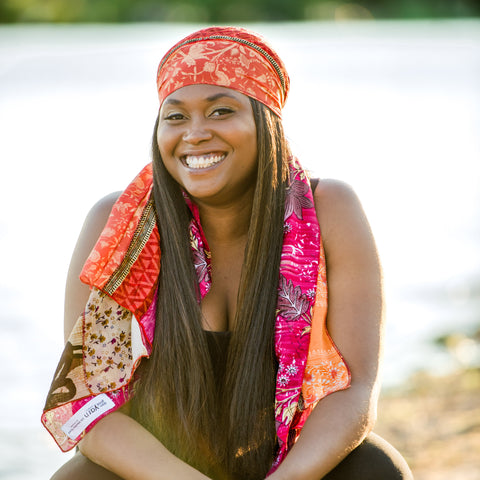 A smiling black woman is sitting on the beach, wearing a pink and yellow medley scarf as a headband on her head.