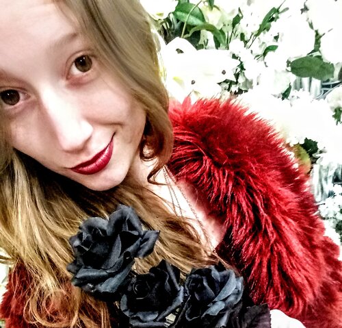 Close up of the author, Michaela Matthews wearing red lipstick and a poofy red scarf with white flower arrangement in background.