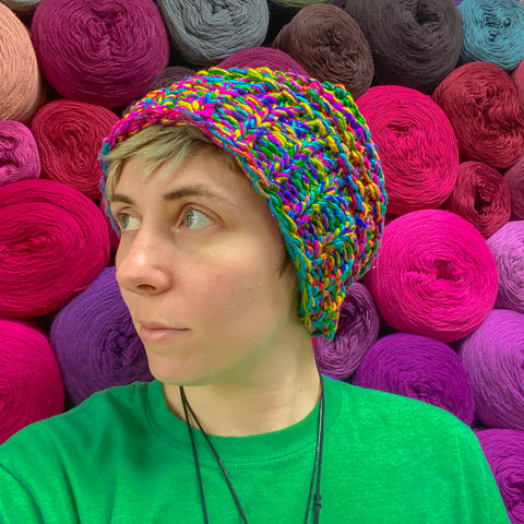 A person with short blonde hair wearing a loom knit beanie, made from watercolors worsted weight silk yarn