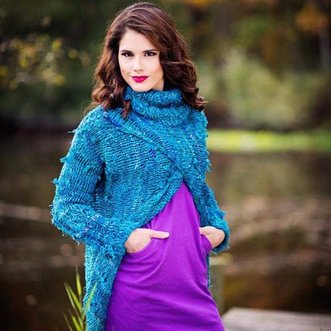 Woman wearing the blue Catwalk Sweater with cowl neck and crossed front standing by greenery