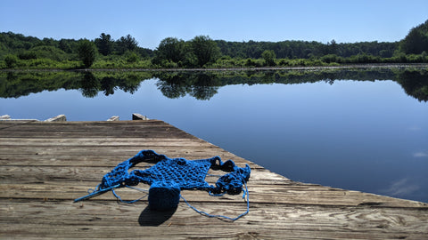 Blue crochet work in progress sitting on a wood dock in front of a river which reflects the blue skies and trees across the way.