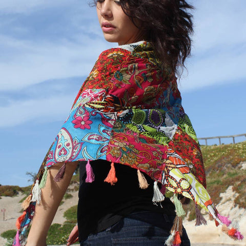 A petite woman with short dark hait and a black tank top is sporting one of our multicolored patchwork scarf with tassels. The woman's body is facing away from the camera and the scarf is blowing in the wind.