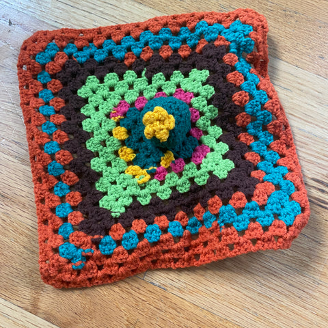 The finished colorful granny square and a crochet hook laying on a wooden floor, finished with the cactus sewn to the center