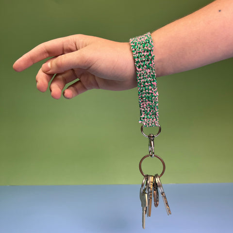 A hand is being held out, with a small key lanyard around their wrist, made from green and pink twist sport weight yarn.