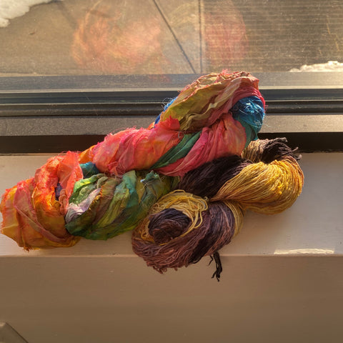 Two dyed skeins of yarn are resting on a windowsill. One skein is yellow and purple, the larger skein is rainbow