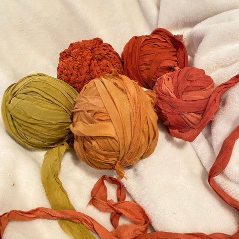 A small orange braided crochet pumpkin and four balls of warm orange and yellow chiffon ribbon yarn are laying on a white blanket.