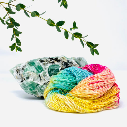 A nest of yellow, green, and pink lace weight silk yarn is sitting in a white room, in front of a large geode with some leafy green plants coming from the top of the image.