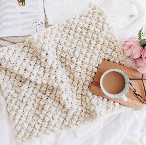 Darn Good Yarn's Raspberry Dreams Baby Blanket on a white sheet with a coffee cup on the right side of the photo.