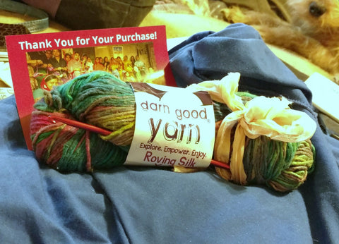 Multicolored skein of yarn, red crochet hook, a photo of female artisans with text that reads 'Thank you for your purchase!' all sitting on a blue fabric surface