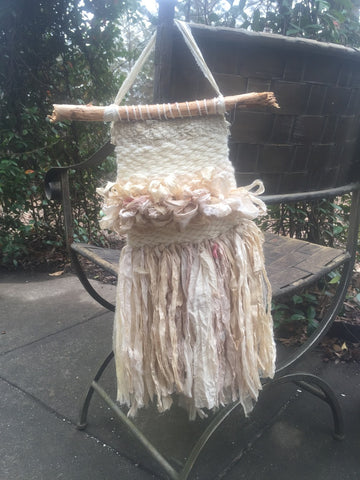 Small white woven wall hanging hung from a metal outdoor chair sitting on top of concrete in front of greenery