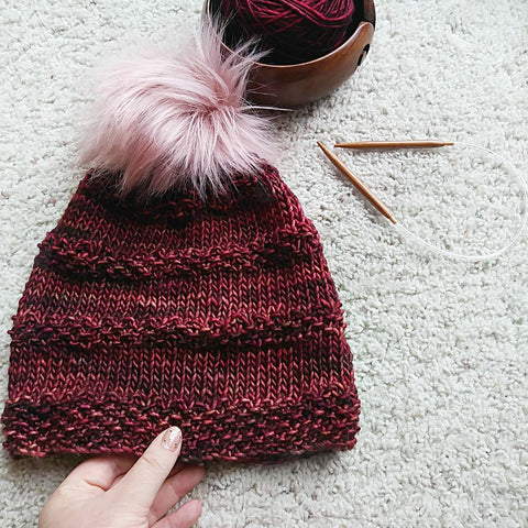 A pom-pom beanie made of deep red yarn is laying on a white carpet.