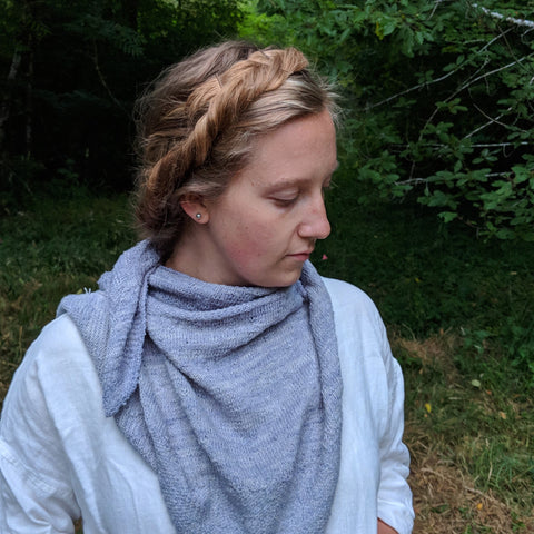 A woman with braided blond hair is wearing a grey handmade shawl over her white long sleeved top outside.