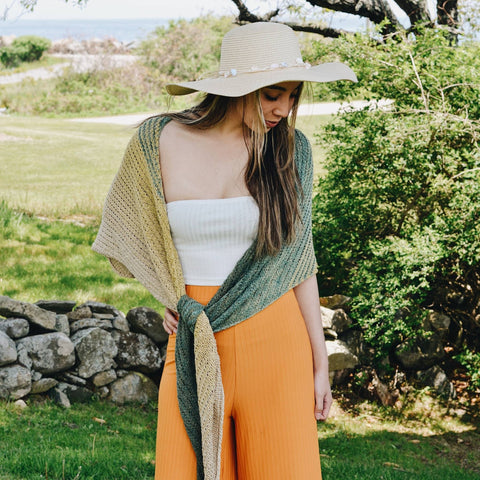 A girl is standing outside, wearing bright orange pants, a white tank top, a white floppy hat, and an ombre green shawl.
