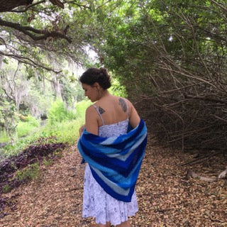 A woman in the woods, facing away from the camera, wrapped in a blue ombre shawl.