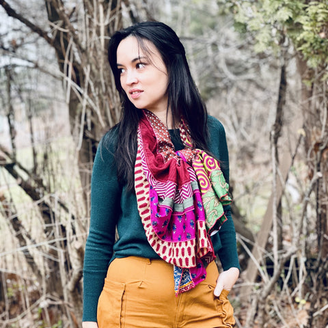 A woman with long black hair is standing in the woods, wearing orange shorts, a teal top, and a brightly colored medley silk sari scarf. 