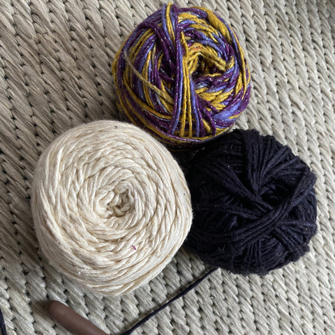 3 cakes of worsted weight yarn are laying on a tan rug. The cakes are black, white, and a mix of purple and yellow sparkle worsted weight yarn.
