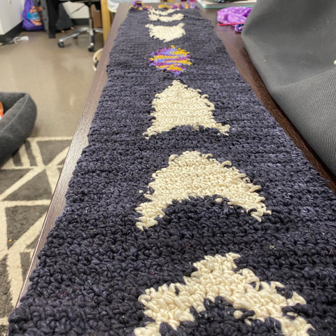 In an office, on a wooden desk, rests the phases of the moon witch's altar runner, made of black, white, and sparkle multicolor yarn. In the center of the runner you can see all the phases of the moon.