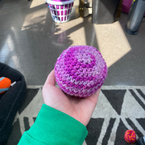 A hand outstretched, holding a medium amigurumi circle made of sparkly ombre pink yarn.