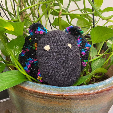 A crochet amigurumi bat made of black and rainbow yarn is sitting in a potted plant, leaning against the green leaves.