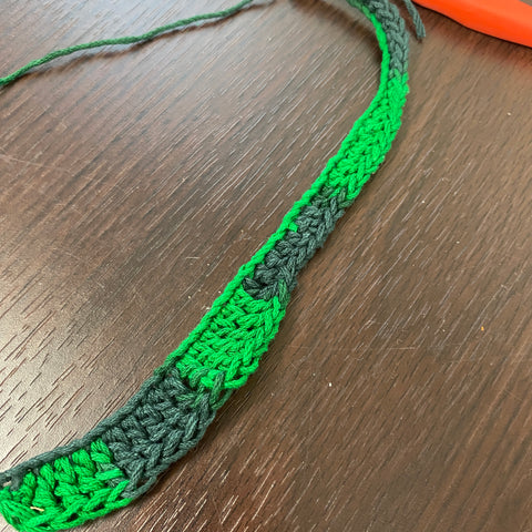 A long green chain of yarn is resting on a wooden desk. The chain is filled with single, double, and triple crochets, creating an increasing row of green yarn.